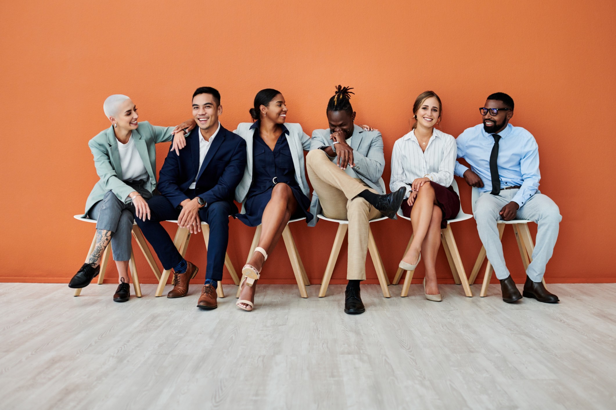 Group of culturally diverse people sitting in chairs against an orange wall.