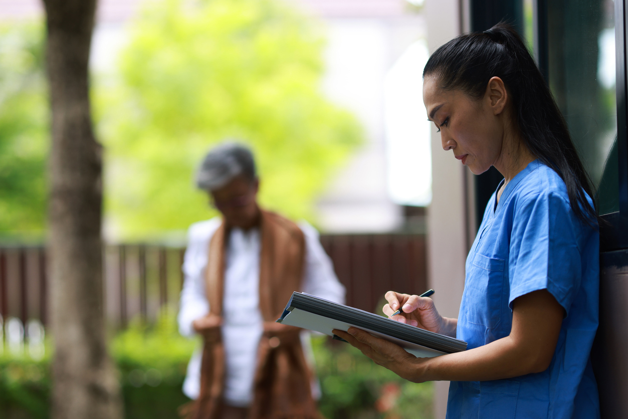 An aged care nurse holding a tablet to do documentation while standing outside. Figure in the background with trees.