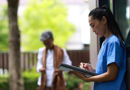 An aged care nurse holding a tablet to do documentation while standing outside. Figure in the background with trees.