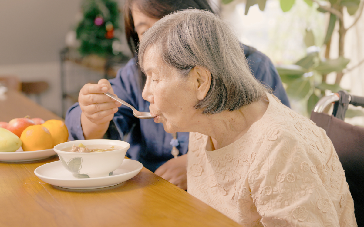 An older person being spoon fed soup by an aged care worker.