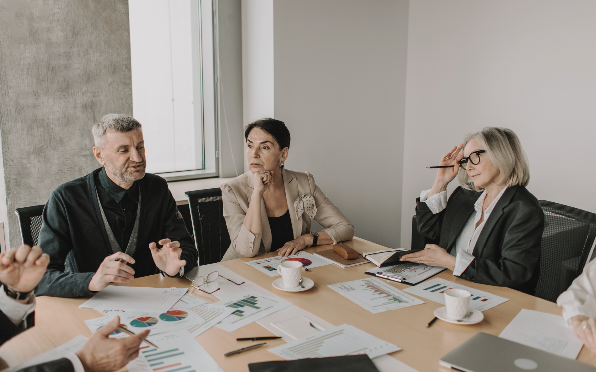A man and two women sit around a table in meeting. There's papers with charts and graphs on them, notebooks and coffee cups. They're in a meeting.